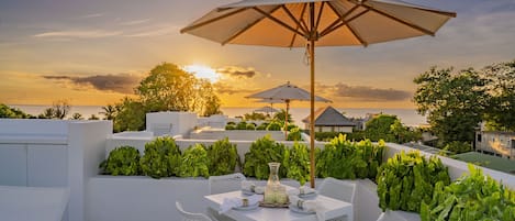An outdoor patio with white furniture, a table set for dining, and a large umbrella providing shade.