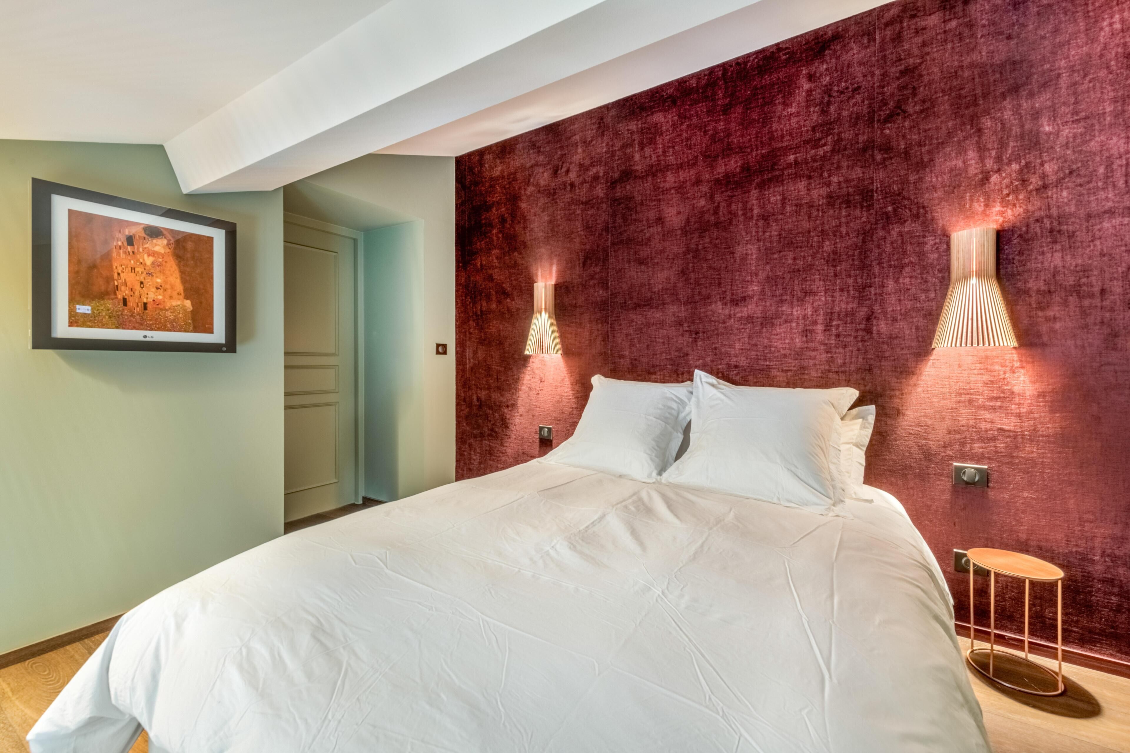 A large bed with white linens, a red textured wall, and two wall-mounted light fixtures.