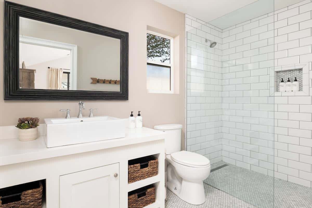 A bathroom with a white sink, mirror, and toilet. There is a shower area with white tiles and a small window.