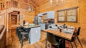 A wooden cabin interior with a kitchen, dining area, and a staircase leading to an upper level.
