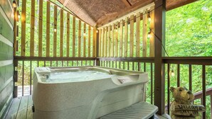 A hot tub on a wooden deck with a 'Welcome' bear statue, surrounded by vertical wooden slats and string lights.