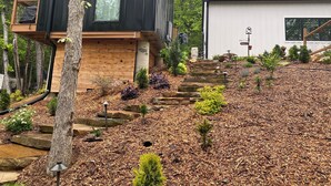 Ein modernes Haus mit einer Holzdiele, einer Steintreppe und einem Garten mit Mulch und kleinen Pflanzen.