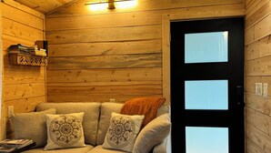 Ein gemütlicher Raum mit Holzvertäfelung, ein beiges Sofa mit gemusterten Kissen, eine schwarze Tür mit Sandstrahlglaseinsätzen und eine kleine Regal mit Büchern.