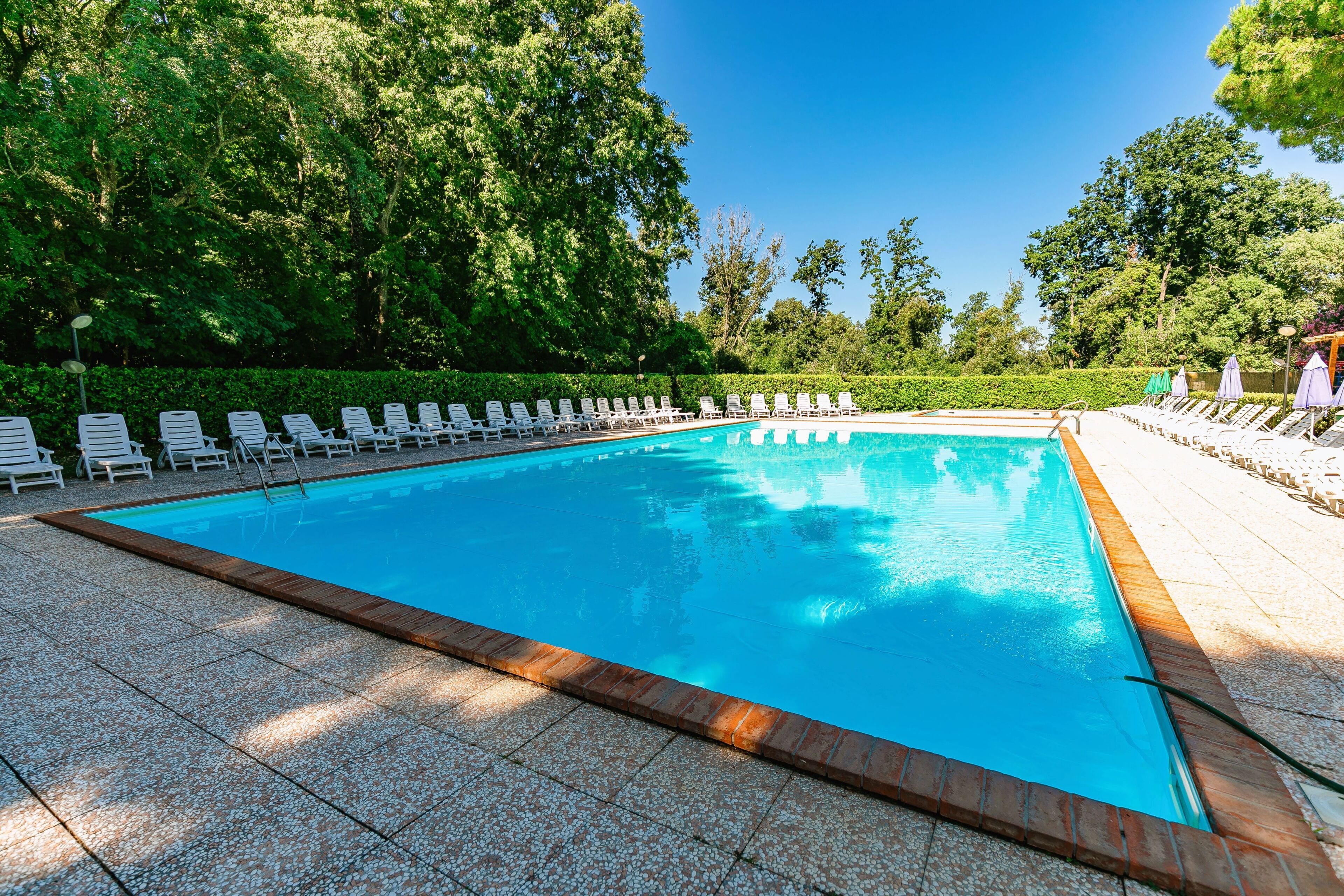 Une piscine rectangulaire, bordée d’une terrasse carrelée, entourée de chaises longues blanches et de parasols, avec des arbres en arrière-plan.