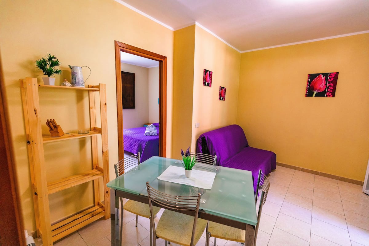 Un pequeño comedor con una mesa de vidrio y sillas metálicas, una estantería de madera y un sofá púrpura al fondo.