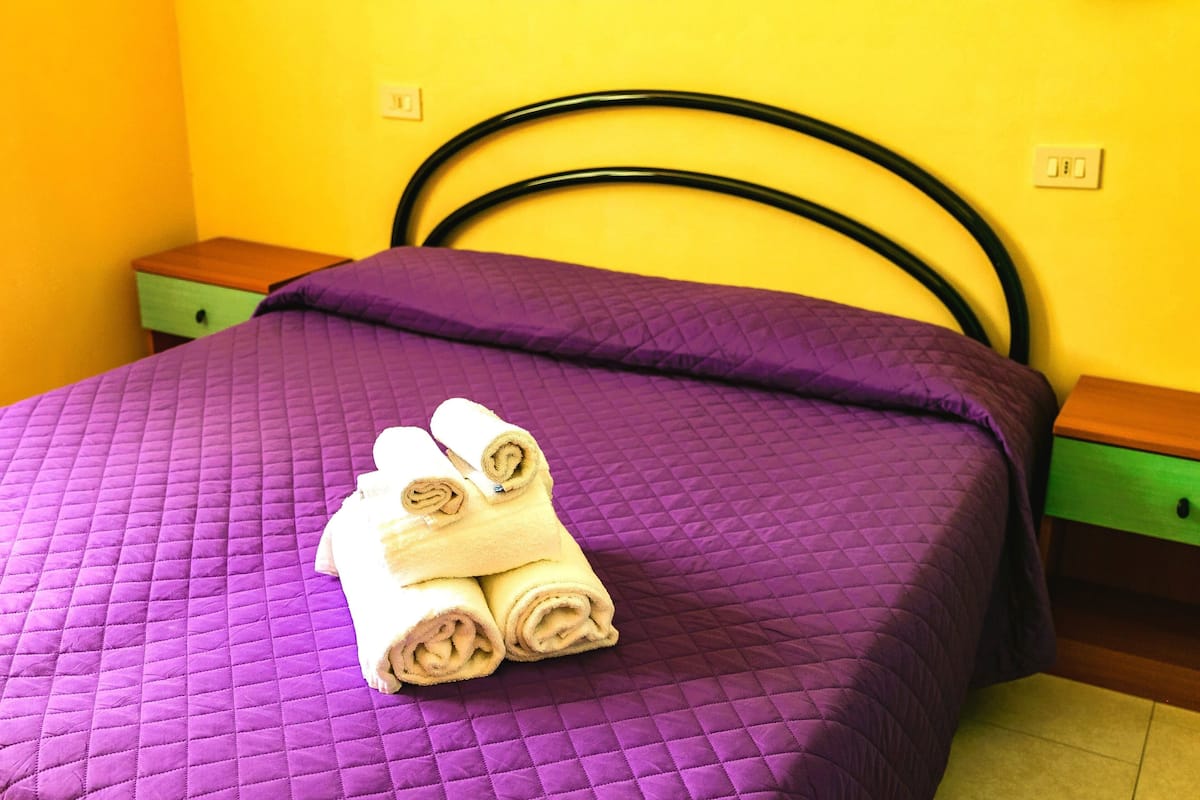 Una cama con una colcha acolchada morada y toallas enrolladas encima.