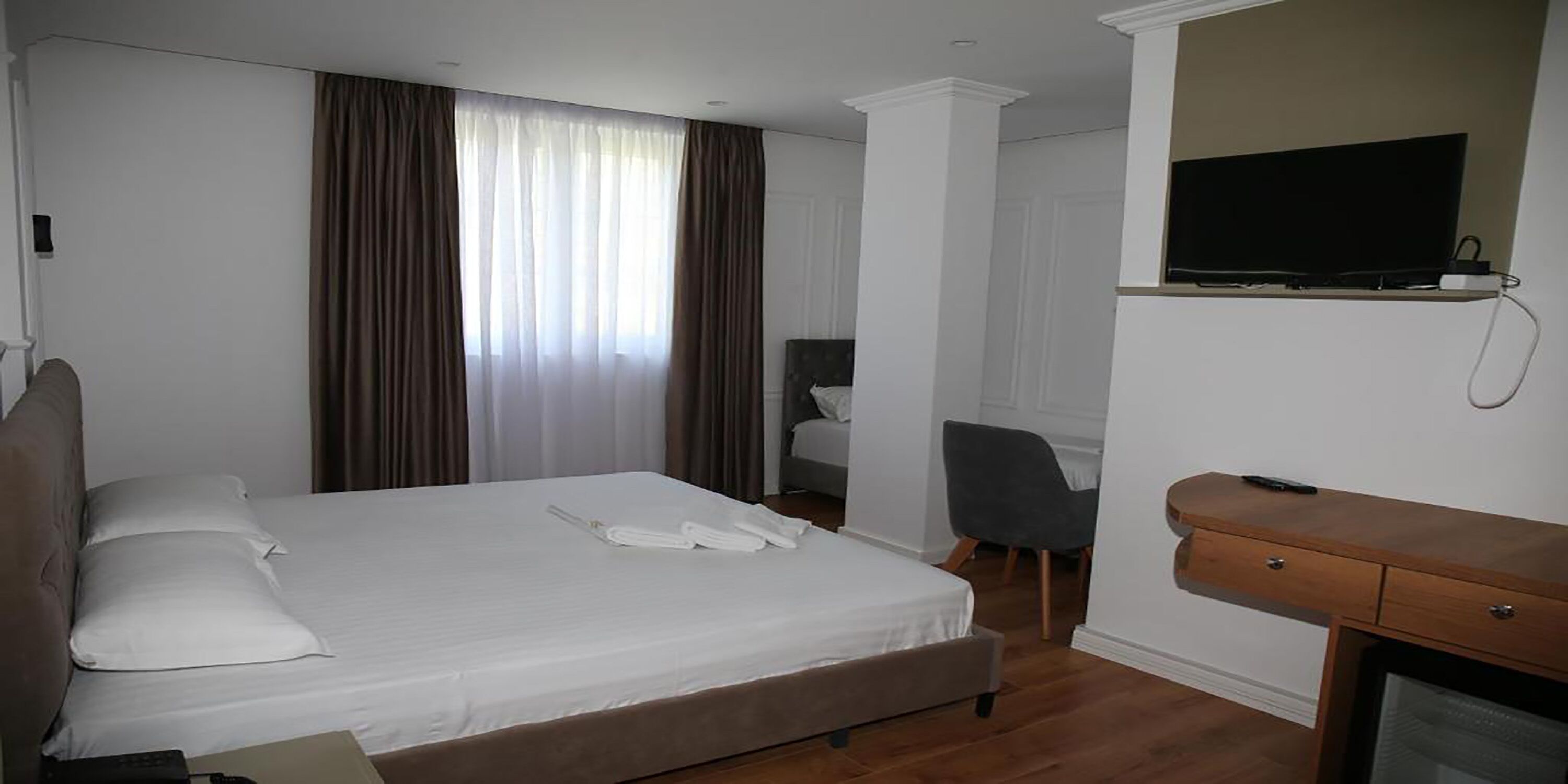 Una habitación de hotel con cama, escritorio, silla, televisor y ventana con cortinas.