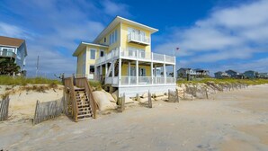 Una casa de playa de varios pisos con un balcón, ubicada en una playa de arena con una cerca de madera y escaleras que conducen a la entrada.