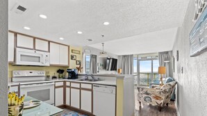 A modern kitchen with white cabinets, a microwave, and a dishwasher. There is a dining area with a patterned rug and a chair. - Bay Watch 632 - Multiple Pools, Hot Tubs, & Lazy River (North Myrtle Beach)