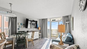 A modern living room with a dining area, a sofa with decorative pillows, a TV mounted on the wall, and a balcony with a view. - Bay Watch 632 - Multiple Pools, Hot Tubs, & Lazy River (North Myrtle Beach)