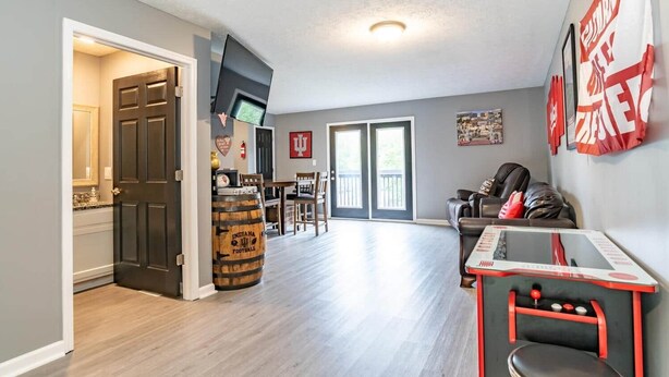 A spacious living area with a dining table, a sofa, and a TV mounted on the wall. There is a large window with a view of the outdoors. - Memorial Field Lodge - 4 Bd Townhome by IU Sports (Bloomington)
