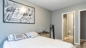 Een slaapkamer met een groot bed, een nachtkastje met een lamp, een spiegel en een lijst met een afbeelding aan de muur.