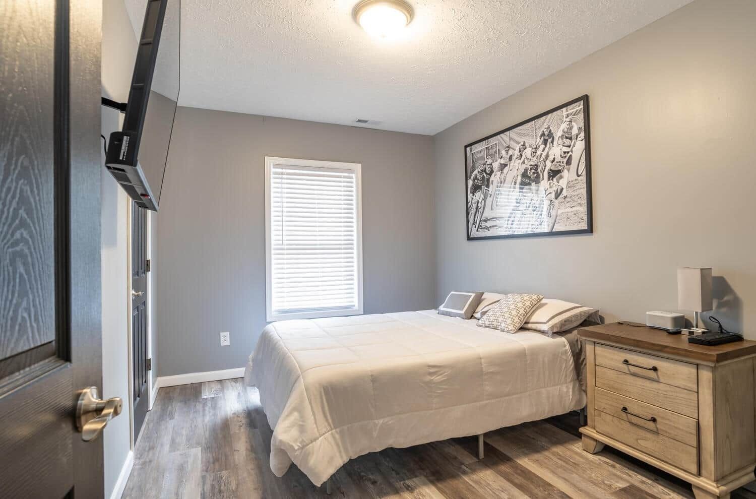 A bedroom with a bed, a nightstand, a dresser, a window with blinds, and a ceiling light.