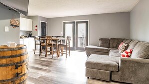 Een moderne woonkamer met een grijze bank, een houten eettafel en stoelen. Een houten vat is in de voorgrond zichtbaar.