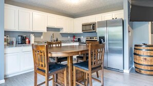A modern kitchen with white cabinets, a stainless steel refrigerator, a microwave, and a wooden dining table with chairs. - IU Soccer Hideaway - 4 Bd Townhome by IU Sports (Bloomington)