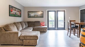A living room with a sectional sofa, a dining table with chairs, and a large window with a view of the outdoors. - IU Soccer Hideaway - 4 Bd Townhome by IU Sports (Bloomington)