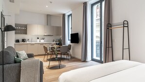 Ein modernes Hotelzimmer mit Bett, einer Couch, einem Esstisch mit Stühlen, einer Küchenzeile und einem Balkon mit Vorhängen.