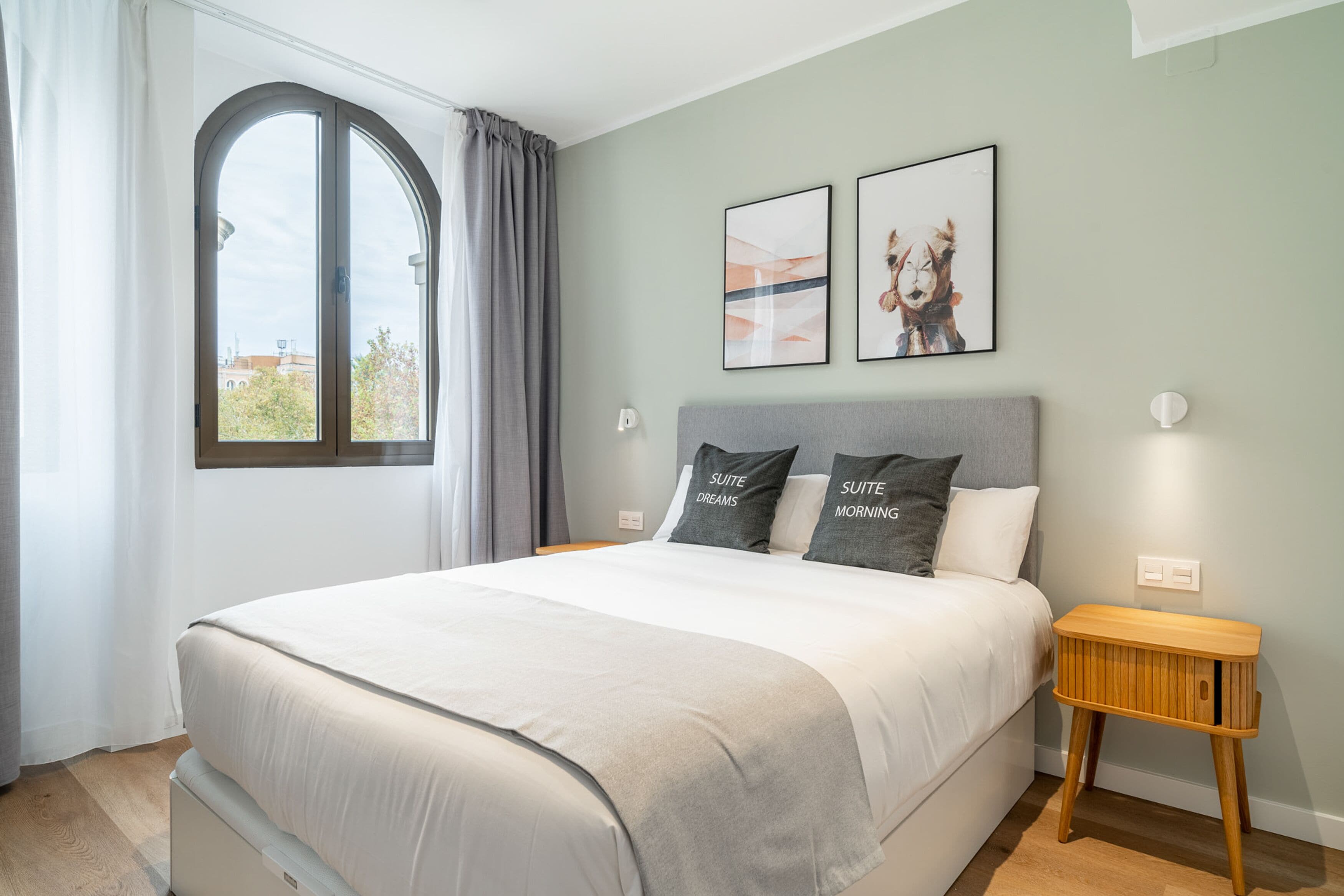 A bedroom with a large bed, a window with curtains, two framed pictures on the wall, and a bedside table with a lamp.