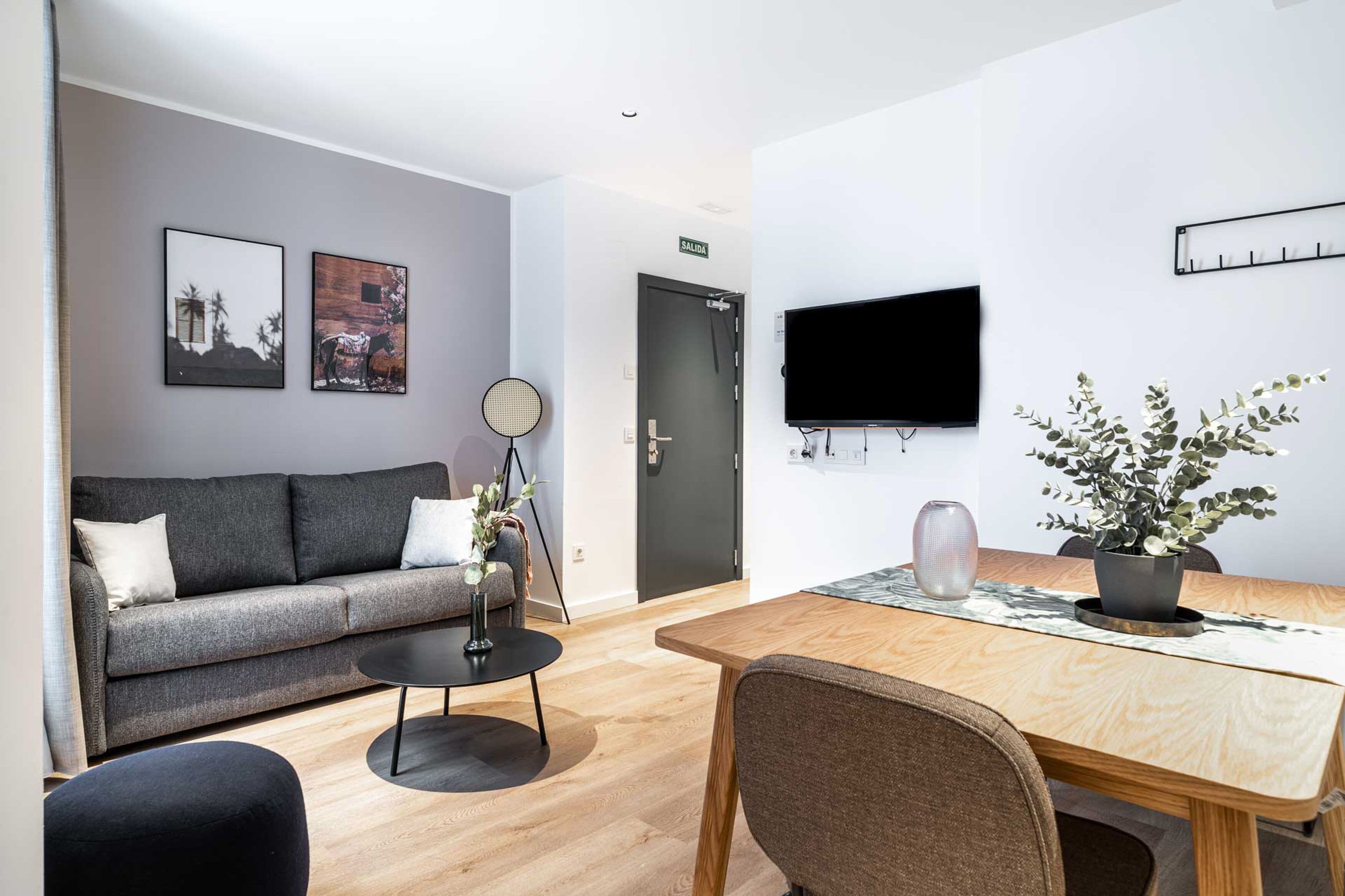Una sala de estar moderna con un sofá gris, una mesa redonda de centro, una mesa de comedor de madera con un jarrón y una planta, y un televisor montado en la pared.