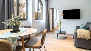 Ein modernes Wohnzimmer mit Essbereich, einem an der Wand befestigten Flachbildfernseher und großen Fenstern mit Vorhängen.