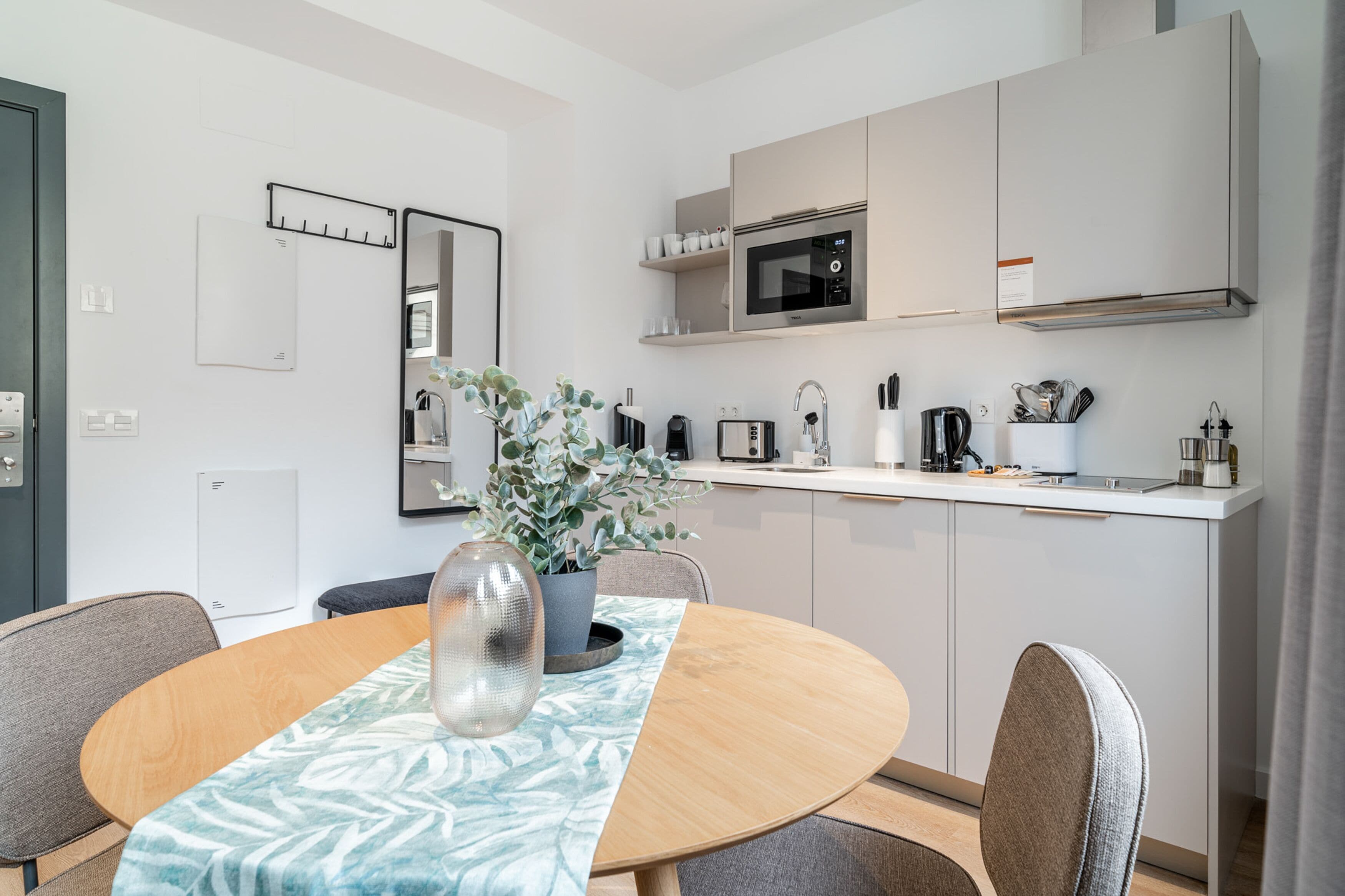 A modern kitchen with a microwave, toaster, and coffee maker. There is a dining table with chairs and a vase with a plant.