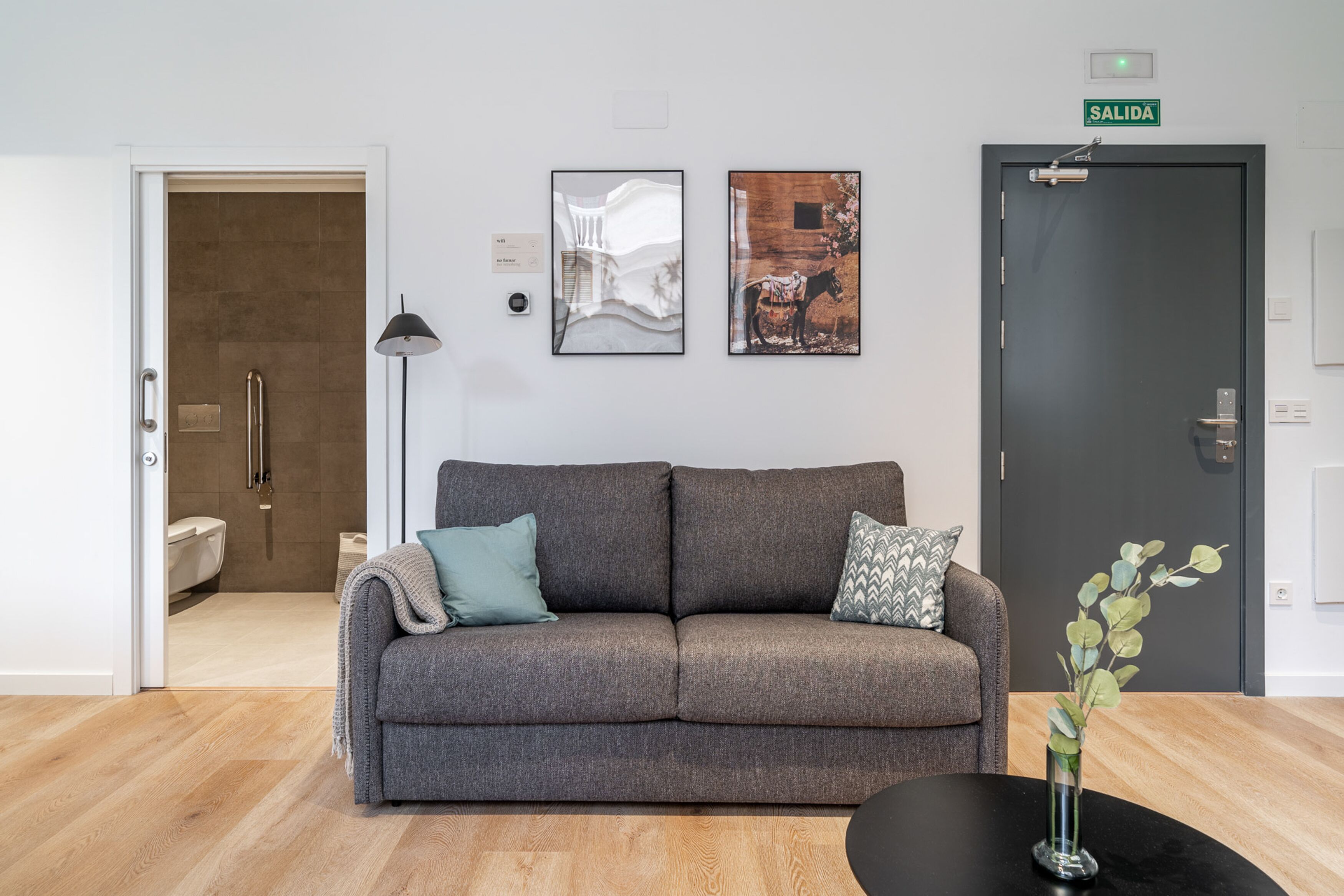 A modern living room with a grey sofa, two pillows, a black coffee table, and a vase with green leaves. There is a door labeled "SALIDA" and a framed picture on the wall.