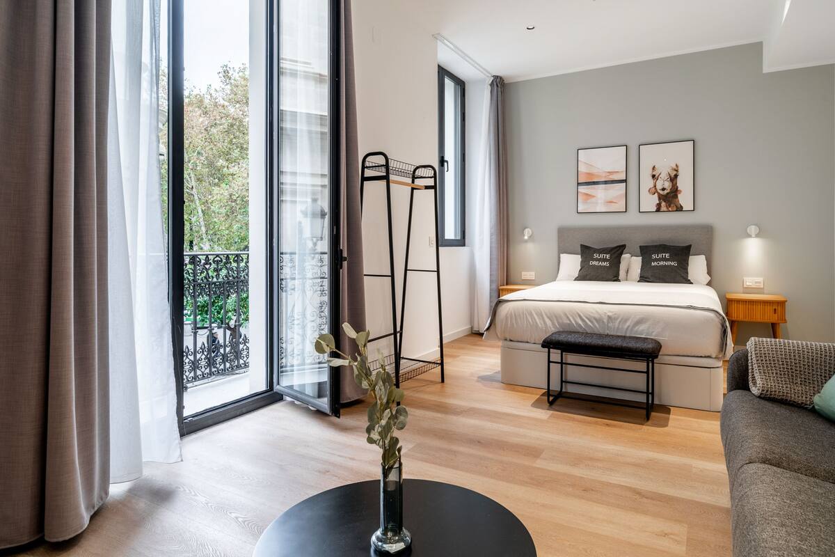 Une chambre d’hôtel moderne avec un grand lit, une table de chevet, un petit banc et une vue sur l’extérieur grâce à de grandes fenêtres.