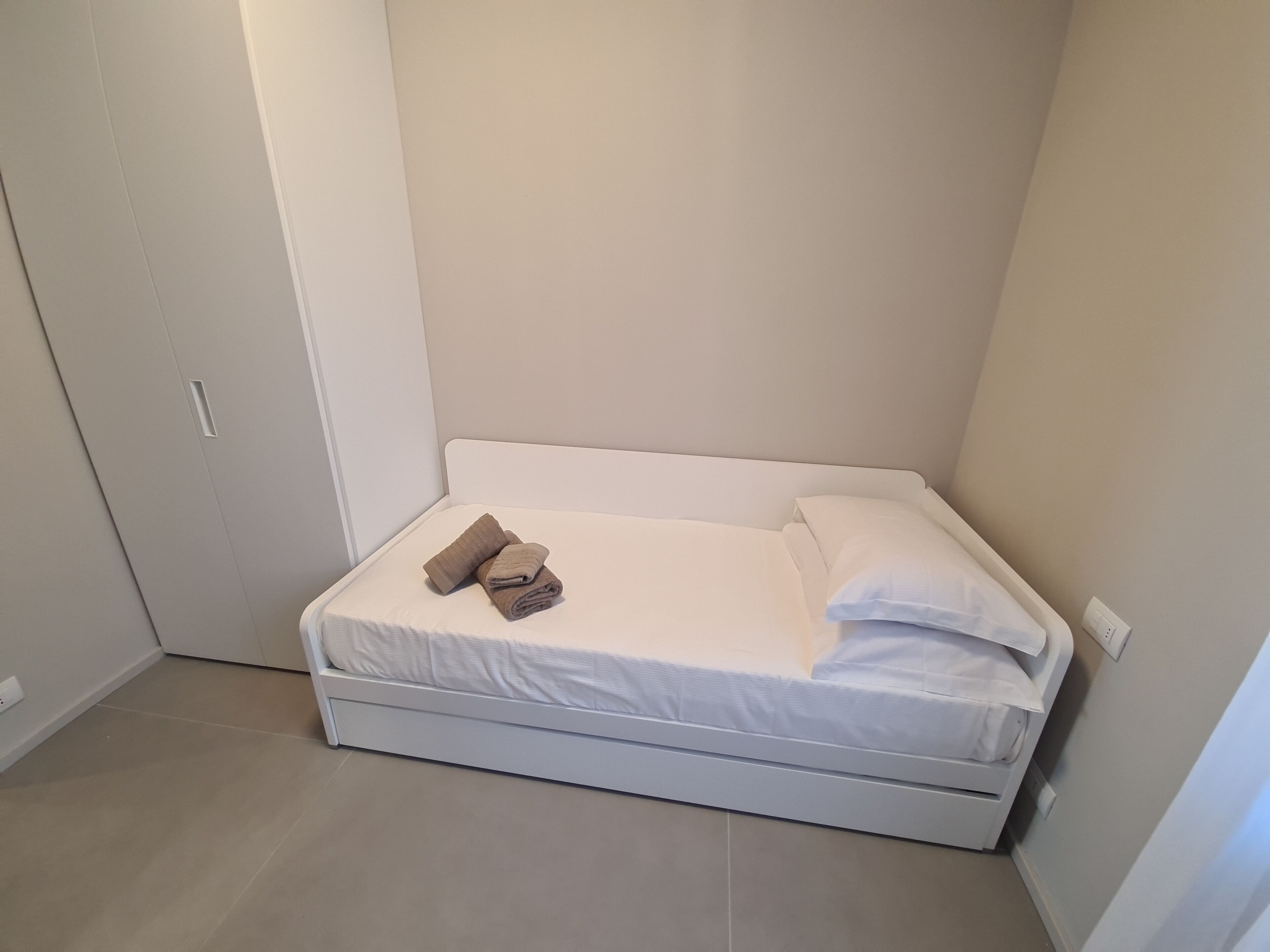 A small, neatly made bed with white bedding and two folded towels in a corner room with white walls and a white door.