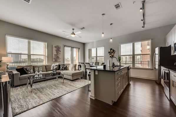 A spacious living room with a sofa, coffee table, and a dining area. There is a kitchen with a large island and a refrigerator.