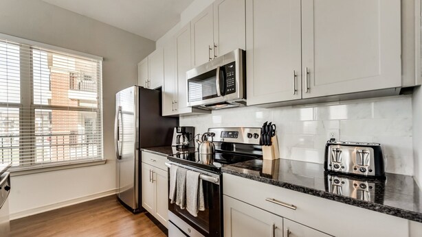A modern kitchen with stainless steel appliances, white cabinetry, a window with blinds, and a tiled backsplash.