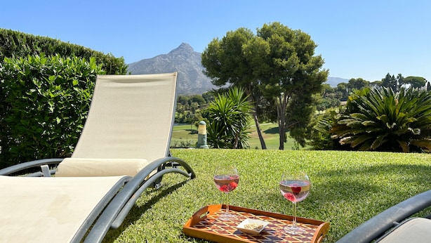 Un espacio al aire libre con césped, una silla de descanso, una bandeja con aperitivos y bebidas, y una vista panorámica de una montaña y árboles.