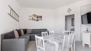 A modern living space with a grey sofa, a dining table with white chairs, and a kitchen area with a TV.