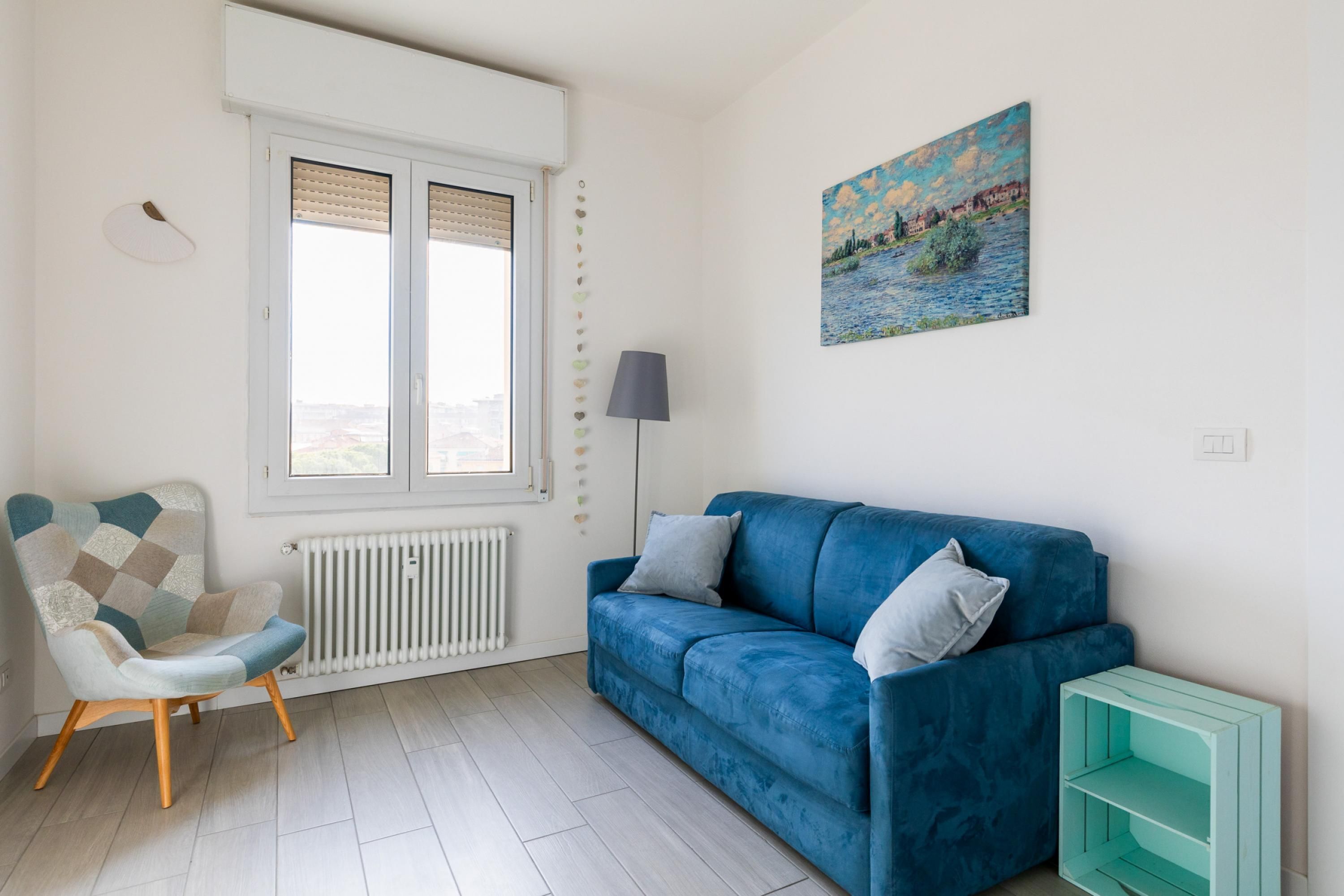 Een kamer met een blauwe bank, een gestreept fauteuil, een radiator, een raam met zonwering, een lamp en een schilderij aan de muur.