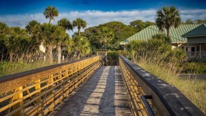 A wooden boardwalk leading to a building with a green roof, surrounded by palm trees and vegetation.