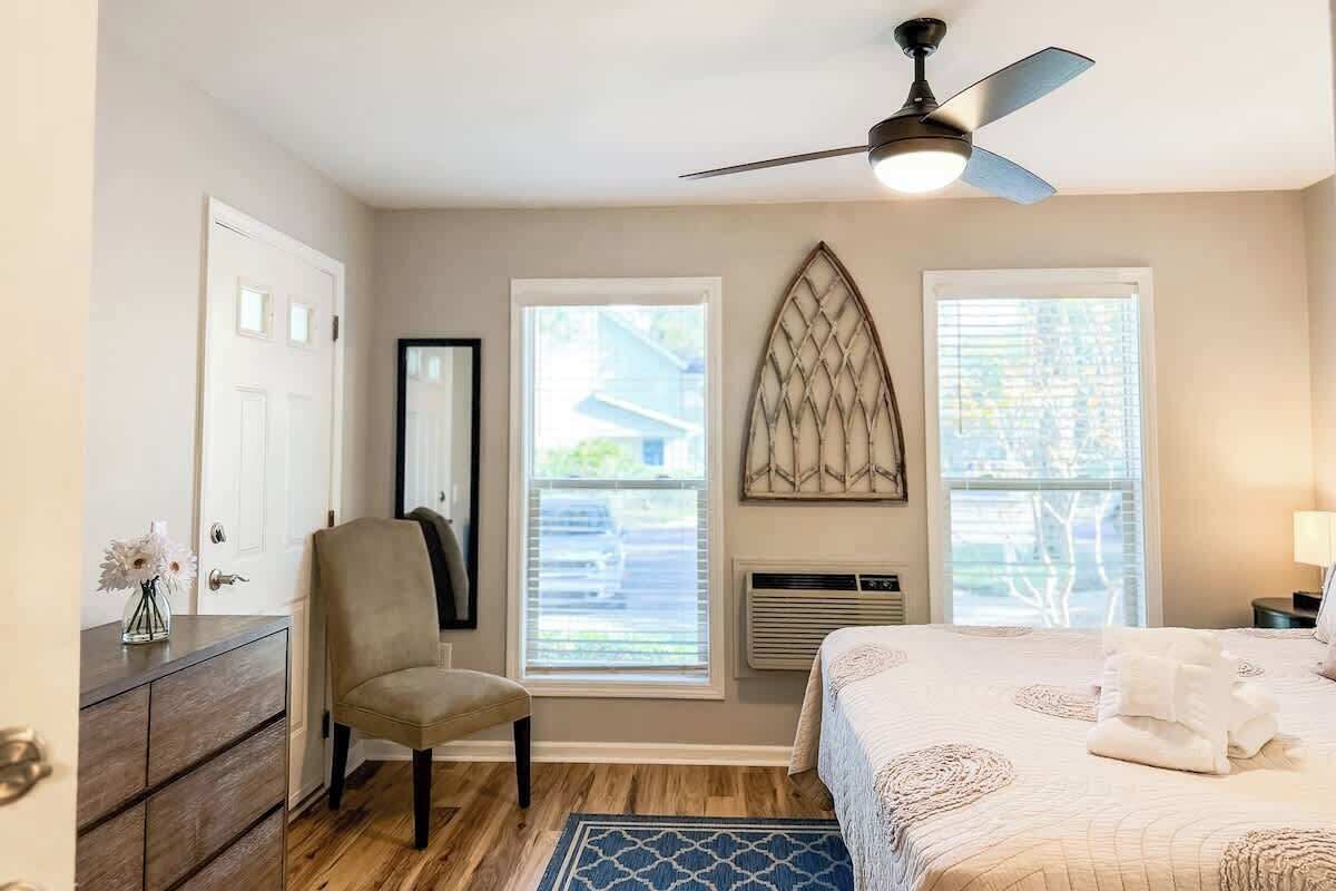 Un dormitorio con una cama, una silla, un tocador, un ventilador de techo y dos ventanas.