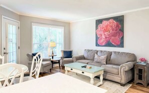 Una sala de estar con un sofá beige, una mesa de centro, una mesa de comedor con sillas, una ventana con persianas y un cuadro grande con motivos florales en la pared.