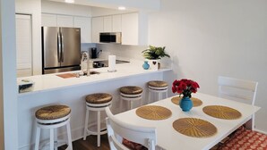 A modern kitchen with white cabinets, stainless steel appliances, and a white dining area with stools and a table. - Oceanfront Queen's Grant Condo "Tide-the-Knot" with Breathtaking Ocean Views! (Topsail Beach)