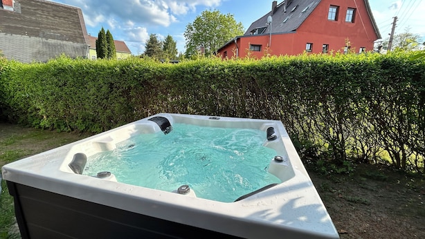 Un jacuzzi dans un jardin, avec une haie, une maison rouge et un ciel dégagé.