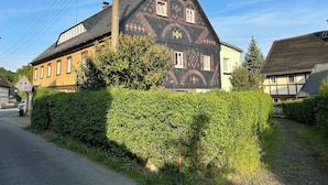 Une maison traditionnelle à deux étages, dotée d’une façade symétrique, d’une cheminée bien visible et d’une haie bien entretenue à l’avant.