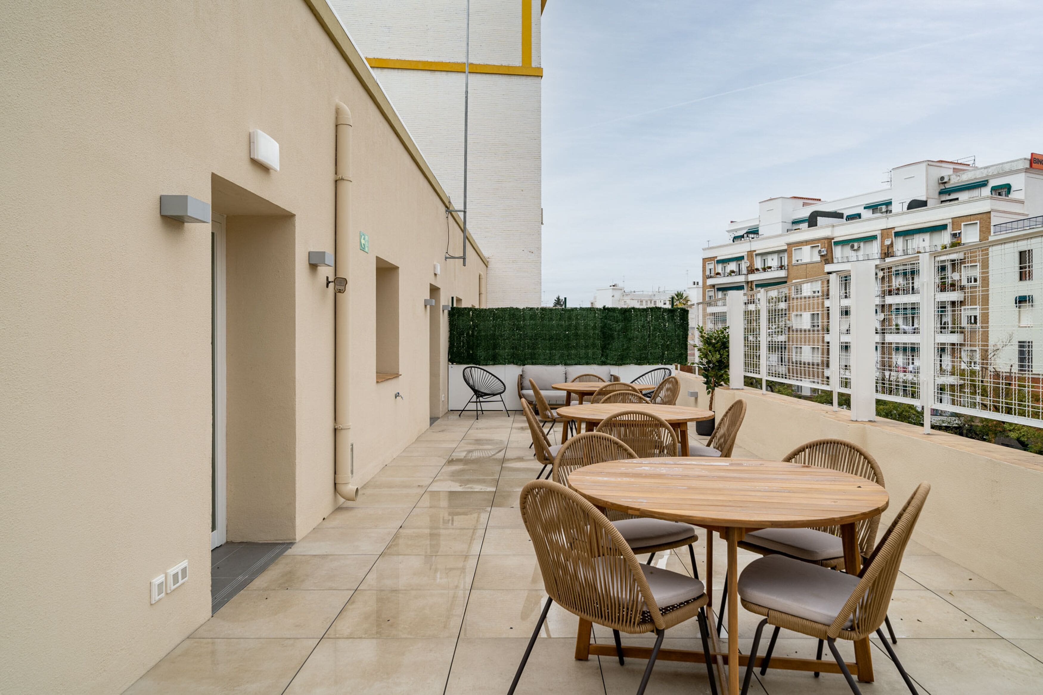 A rooftop terrace with a long table and chairs, a green wall, and a view of other buildings.