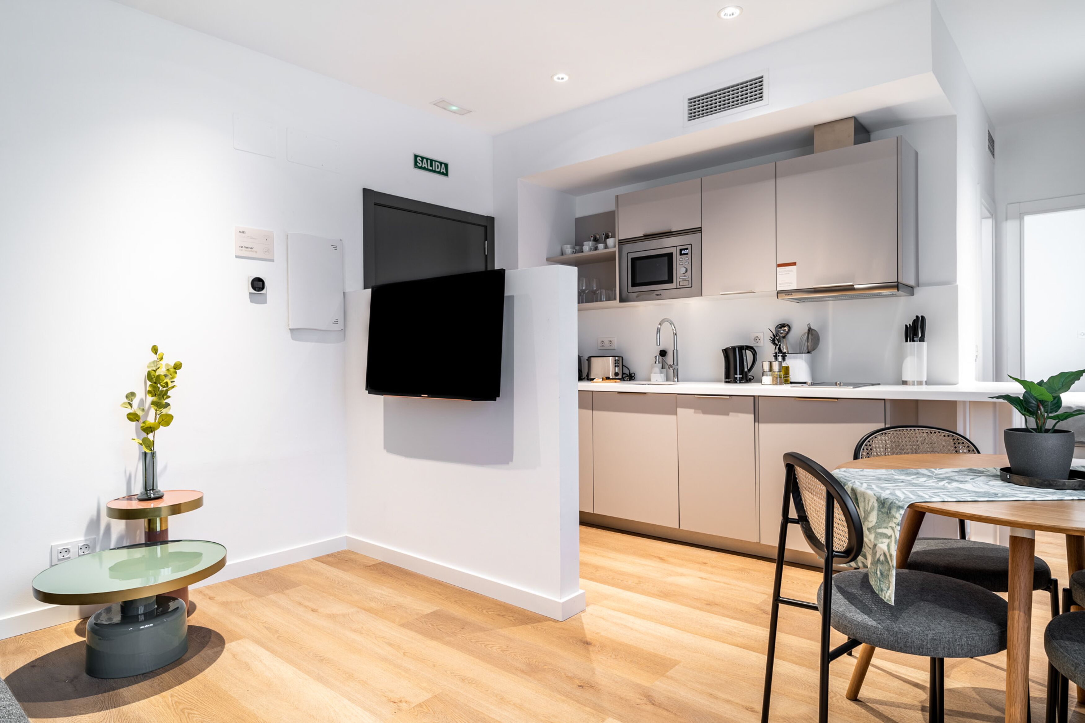A modern kitchen with white cabinets, a microwave, and a sink. There is a dining table with chairs and a potted plant.