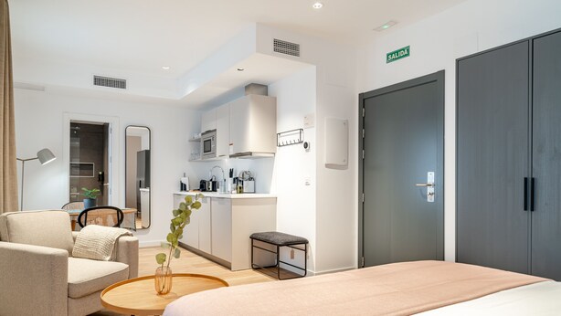 A modern hotel room with a bed, a seating area with a chair and a small table, a kitchenette, and a wardrobe. - Limehome Sevilla Ronda de Capuchinos (Seville)