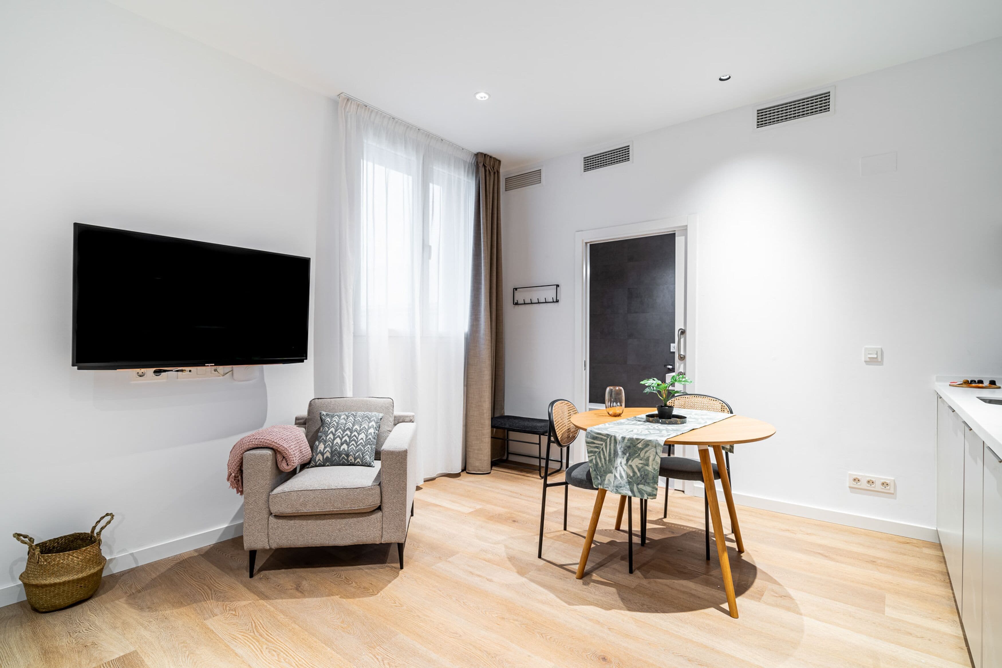 Una sala de estar moderna con un televisor de pantalla plana montado en la pared, un sofá, una mesa de comedor con sillas y una zona de cocina con fregadero y encimera.