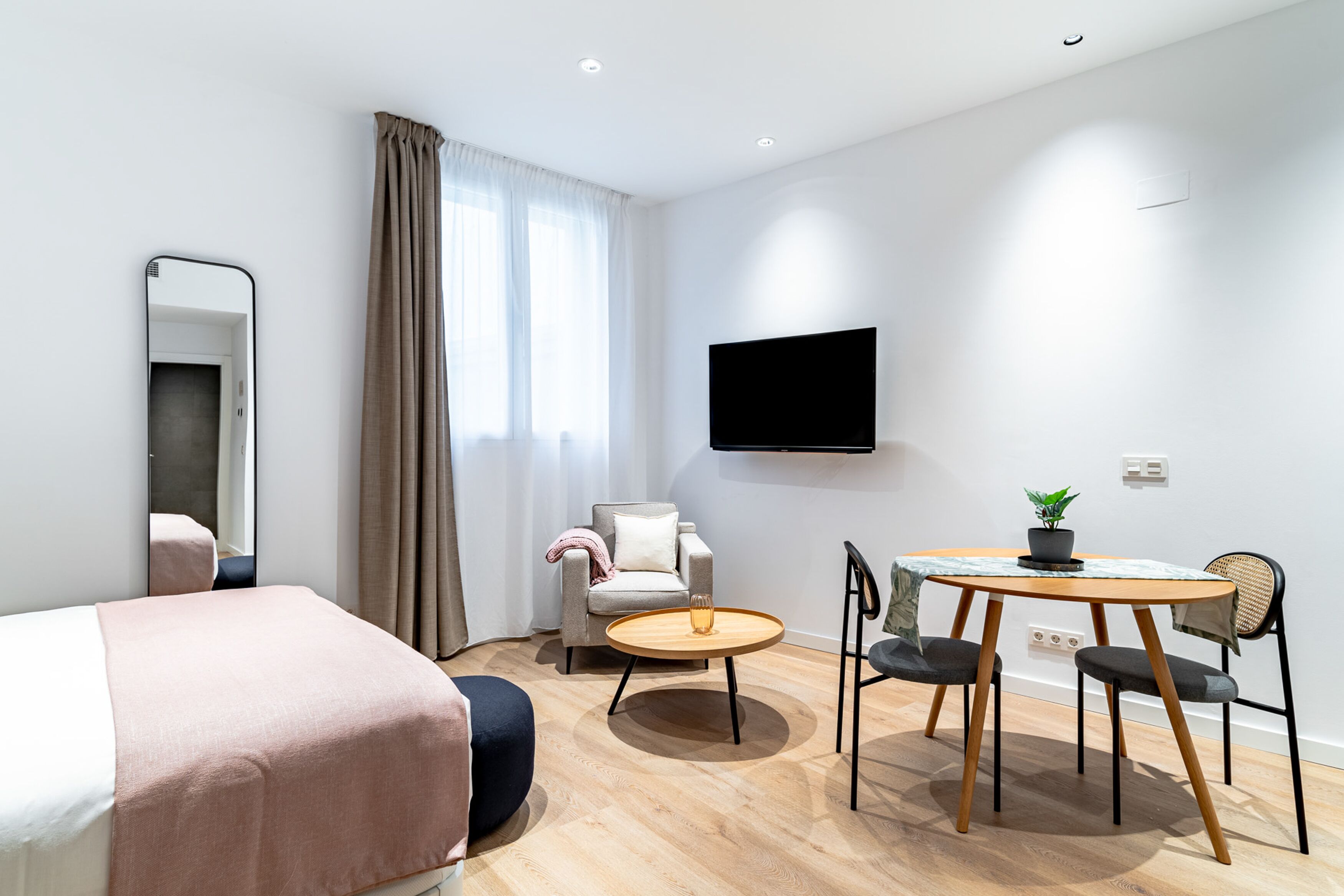 Een moderne hotelkamer met een bed, een bank, een eettafel met stoelen, een aan de muur bevestigde televisie en een raam met gordijnen.