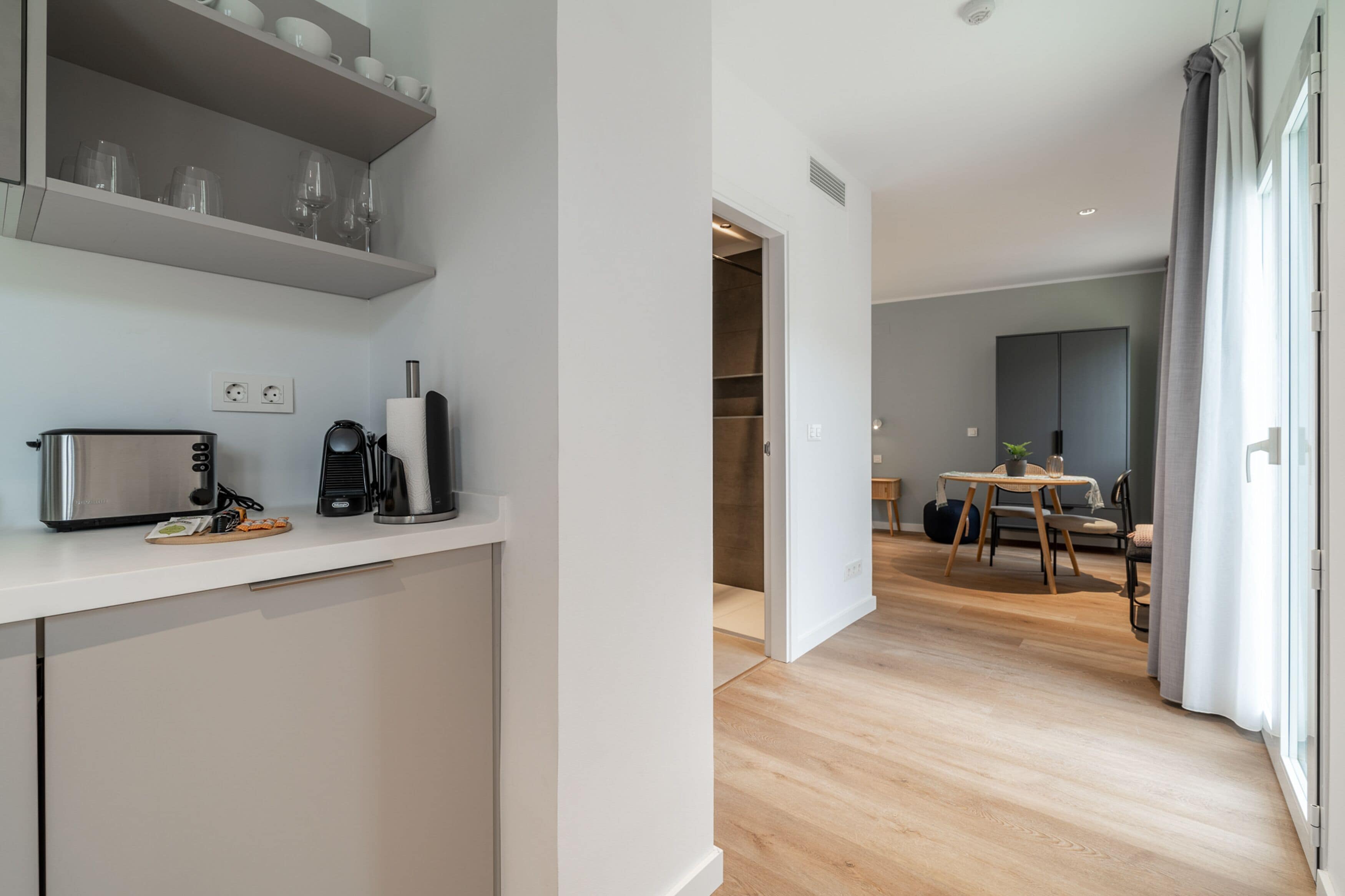 A modern kitchen with white cabinets, a microwave, and a coffee maker. There is a dining area with a table and chairs, and a large window with curtains.