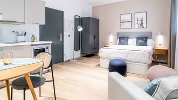 Quarto de hotel moderno com cama, mesas de cabeceira, escrivaninha e cozinha compacta.