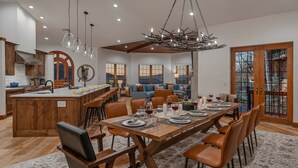 A spacious kitchen with a central island, a dining area with a long wooden table set for six, and a cozy living area with a blue sofa and a view of the mountains.