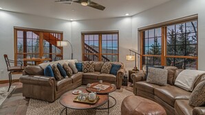 A spacious living room with a large sectional sofa, a coffee table, and a fireplace. There are large windows offering a view of the outdoors.