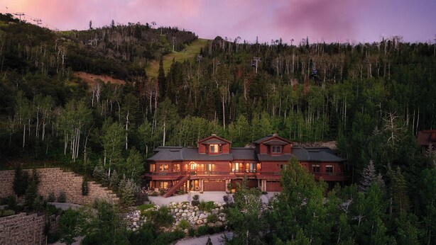 A large, multi-story lodge with a stone foundation, surrounded by dense forest and a ski lift in the background.