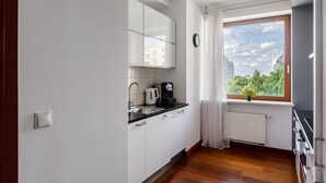 A small kitchen with white cabinets, a black countertop, a sink, a kettle, and a coffee maker. There is a window with a view of buildings and trees. - Bagno (Warsaw)